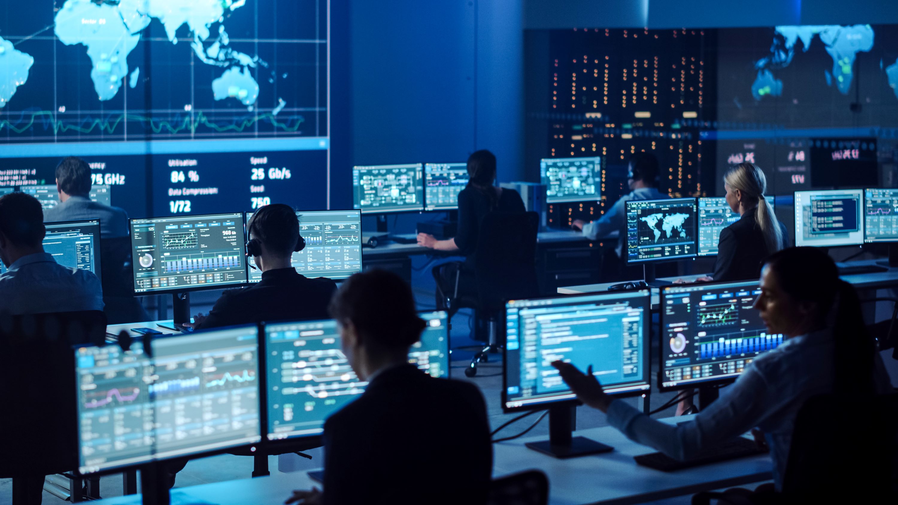 Multiple government workers sitting at desks with dual-monitor computers and within a control center.